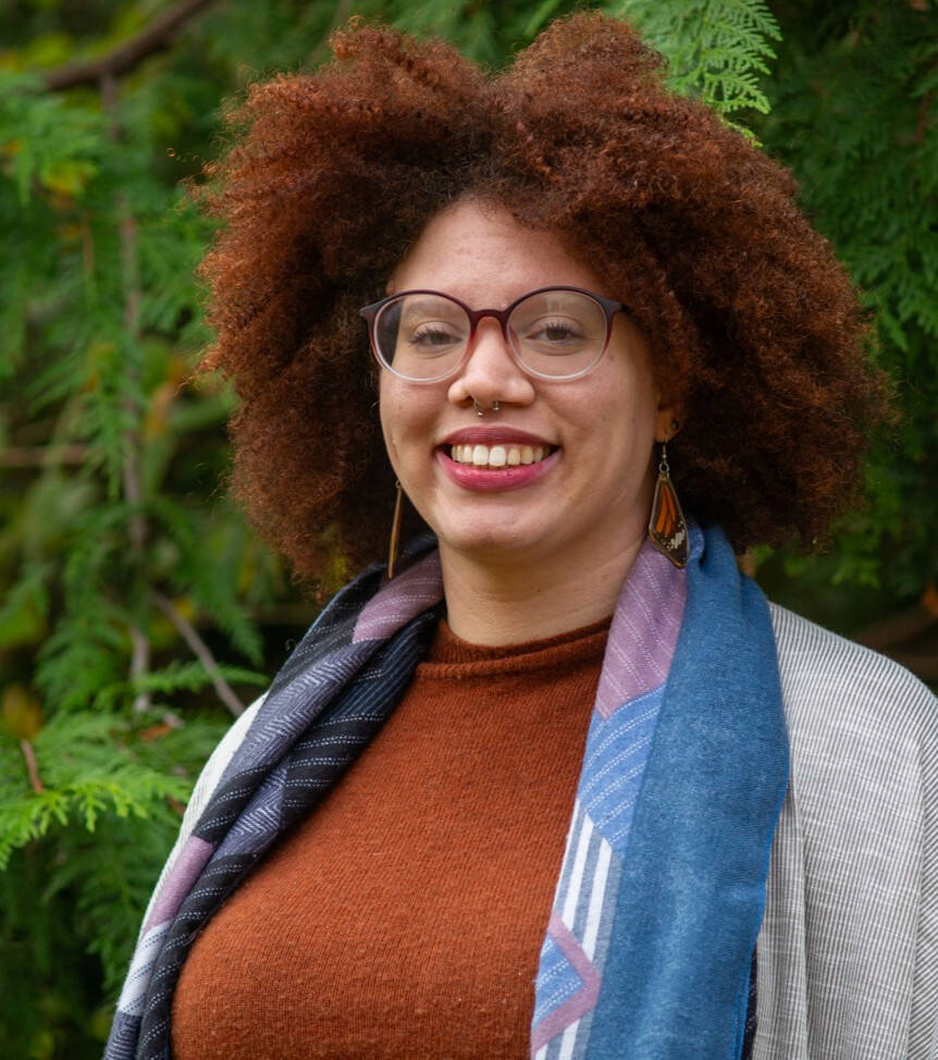 Mixed race woman with light skin tone in front of green shrubs with red curly afro, dark glasses, wearing a warm brown sweater, blue scarf, grey jacket and earrings